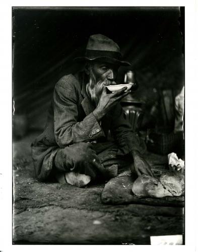 Anciano sorbiendo de un plato con hogaza de pan a sus pies.